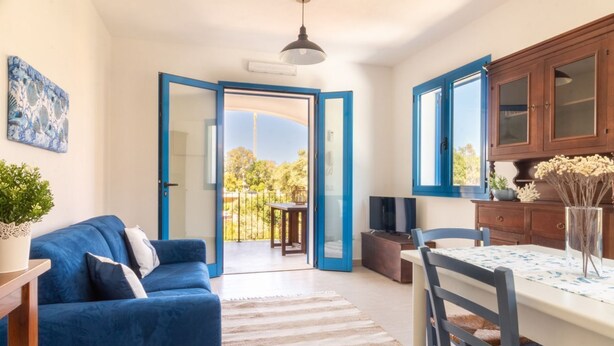 Una sala de estar moderna con un sofá azul, una mesa de comedor con sillas, y una zona de cocina con armarios y un televisor.