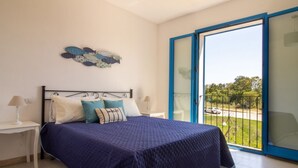 Un dormitorio con cama, mesita de noche, lámpara, una obra de arte en la pared y un amplio ventanal corredero que da a un balcón.