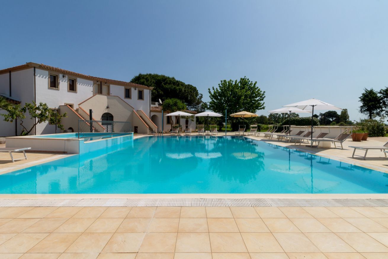 A swimming pool with lounge chairs and umbrellas, surrounded by a building with a tiled roof.