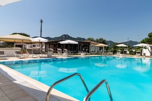 Una piscina con sillas de descanso y sombrillas, rodeada de edificios y montañas.
