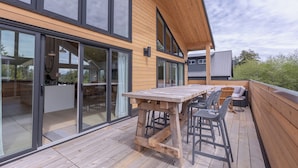 A modern wooden deck with a table and chairs, large sliding glass doors, and a view of the surrounding landscape.