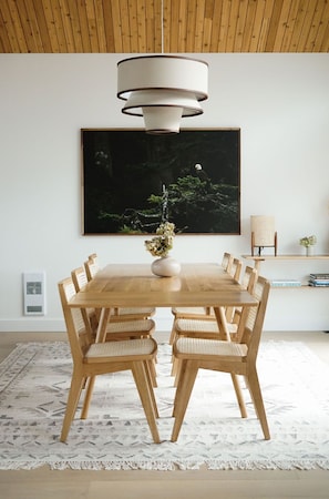 Un coin repas avec une table en bois, six chaises, un vase de fleurs, une grande peinture abstraite, un fauteuil en osier et une bibliothèque remplie de livres.