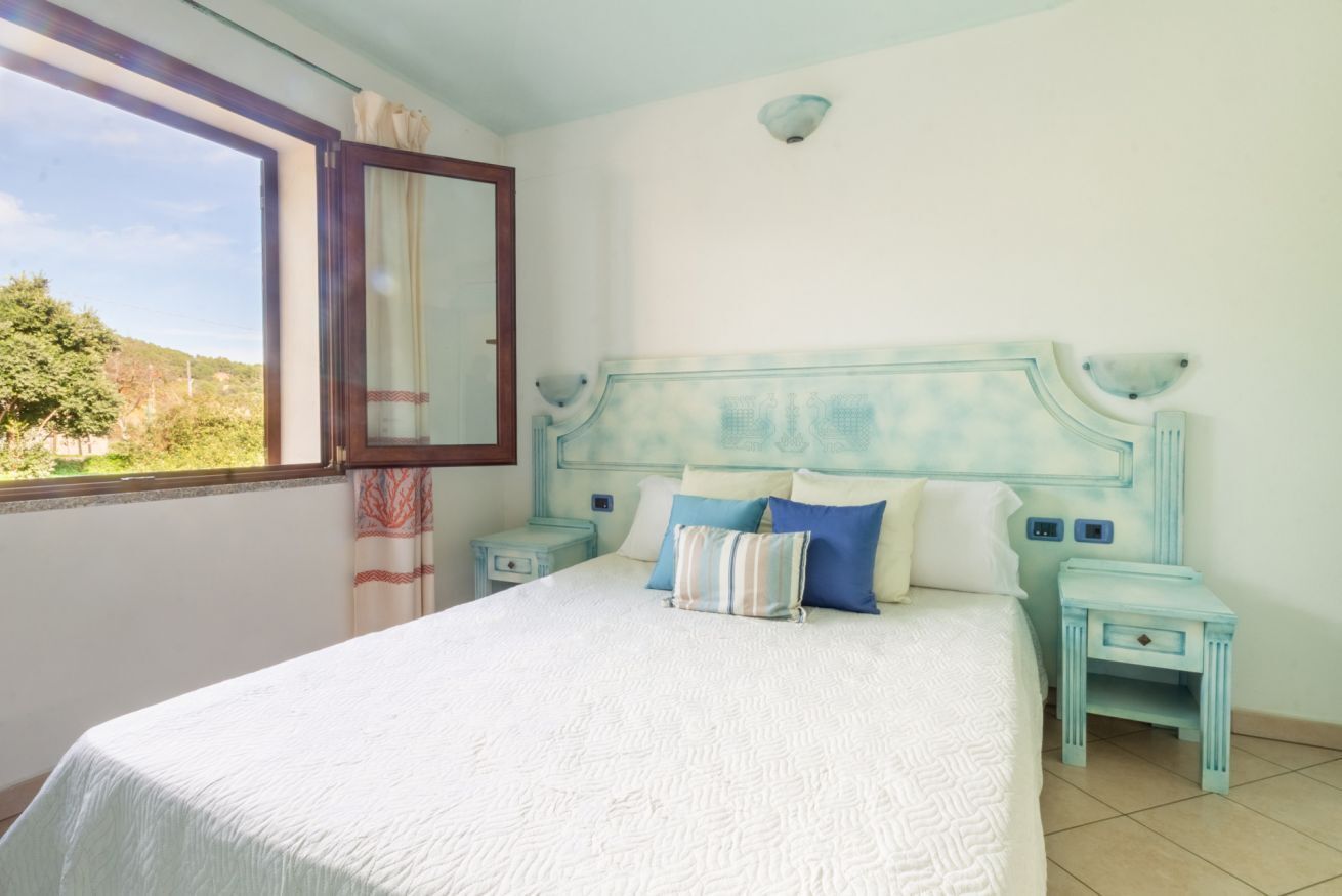 A bedroom with a large bed, a window with curtains, and a view of greenery outside.