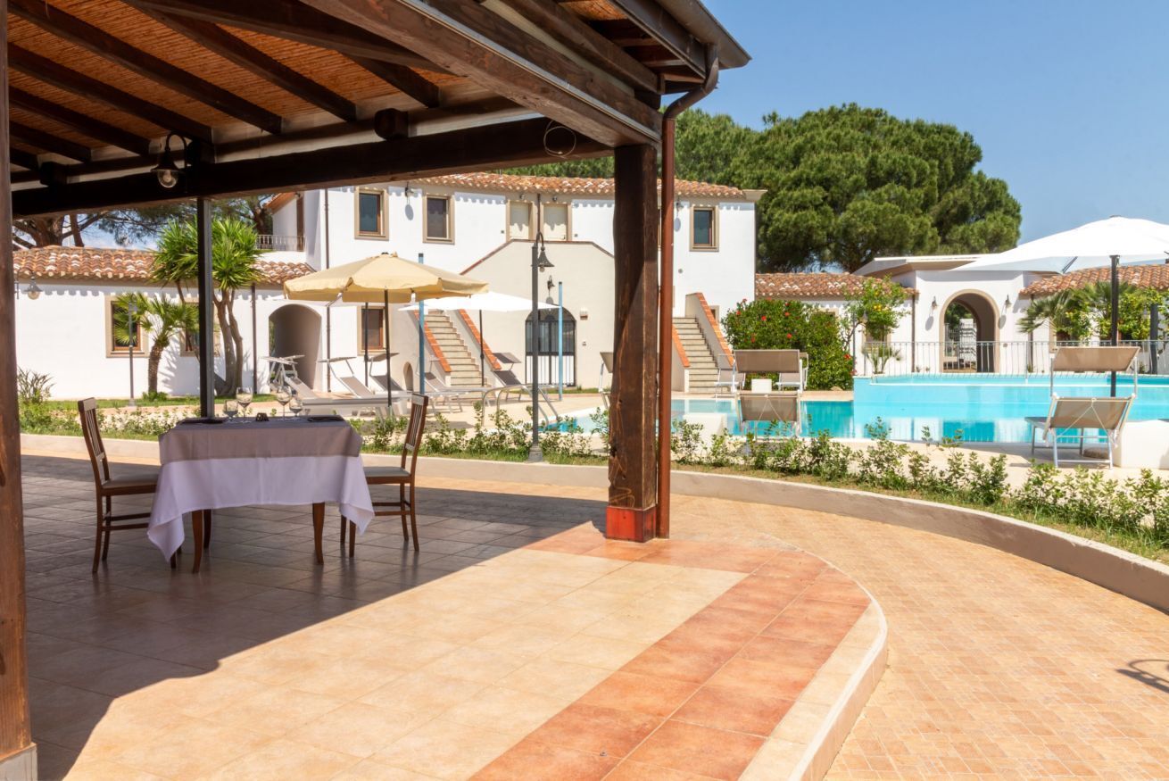 Un'area esterna riparata con tavolo e sedie, una piscina e un edificio con tetto in tegole sullo sfondo.