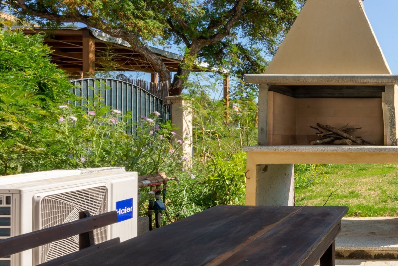 An outdoor grill area with a bench, surrounded by greenery and a fence.