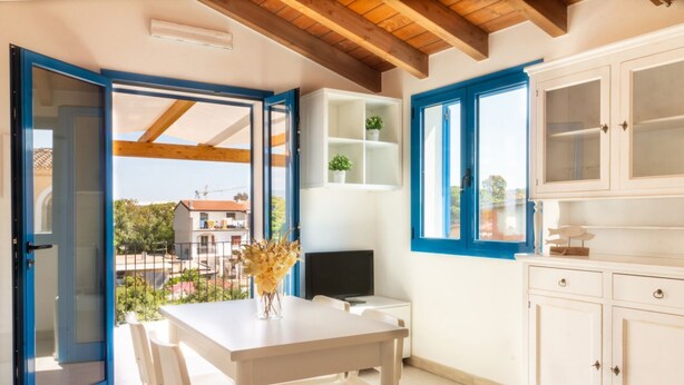 A room with a white table, chairs, a TV, and a cabinet. There is a window with blue trim and a view of the outside. - Sole Apartment by All4sardinia  (Arbatax)