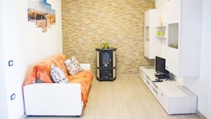 A compact living space with a brick-patterned wall, a white sofa with orange bedding, a small black stove, and a white entertainment unit with a TV.