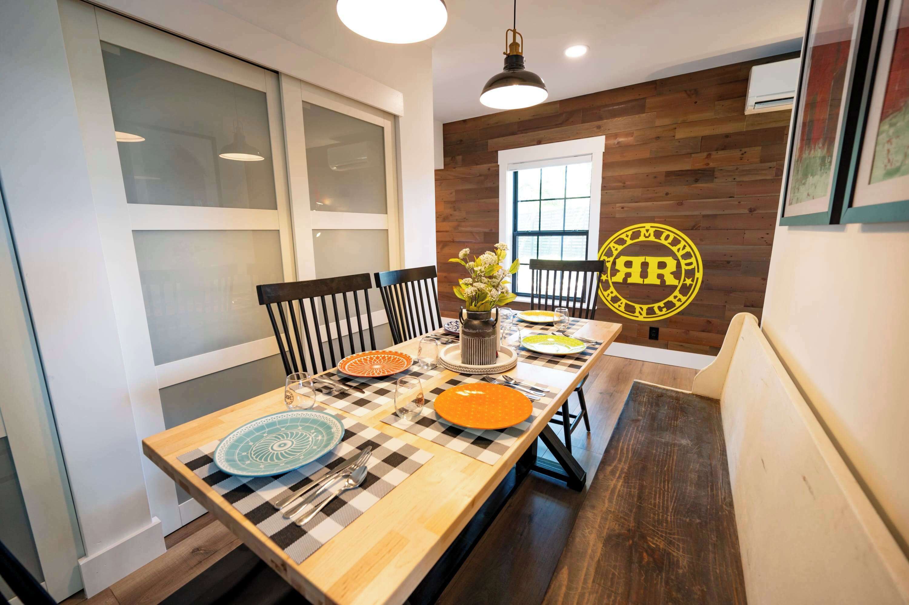 A dining area with a long table set for four, featuring a checkered table runner, a vase with flowers, and a yellow circular logo on the wall.