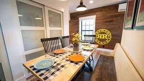 A dining area with a long table set for four, featuring a checkered table runner, a vase with flowers, and a yellow circular logo on the wall.