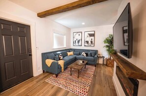 Ein Wohnzimmer mit einer dunklen Holztür, einem blauen Ecksofa, einem Couchtisch aus Holz, einem Teppich mit Muster und einem Kamin mit Fernseher darüber.