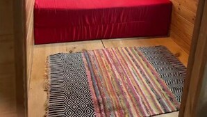 Una habitación de cabaña acogedora con interior de madera, una ventana con persianas, una cama con cubrecama rojo y una alfombra estampada en el suelo.