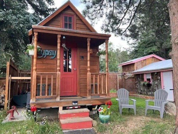 Une cabane en bois avec une porte rouge, un porche et un petit jardin agrémenté de chaises et d’une borne d’incendie.