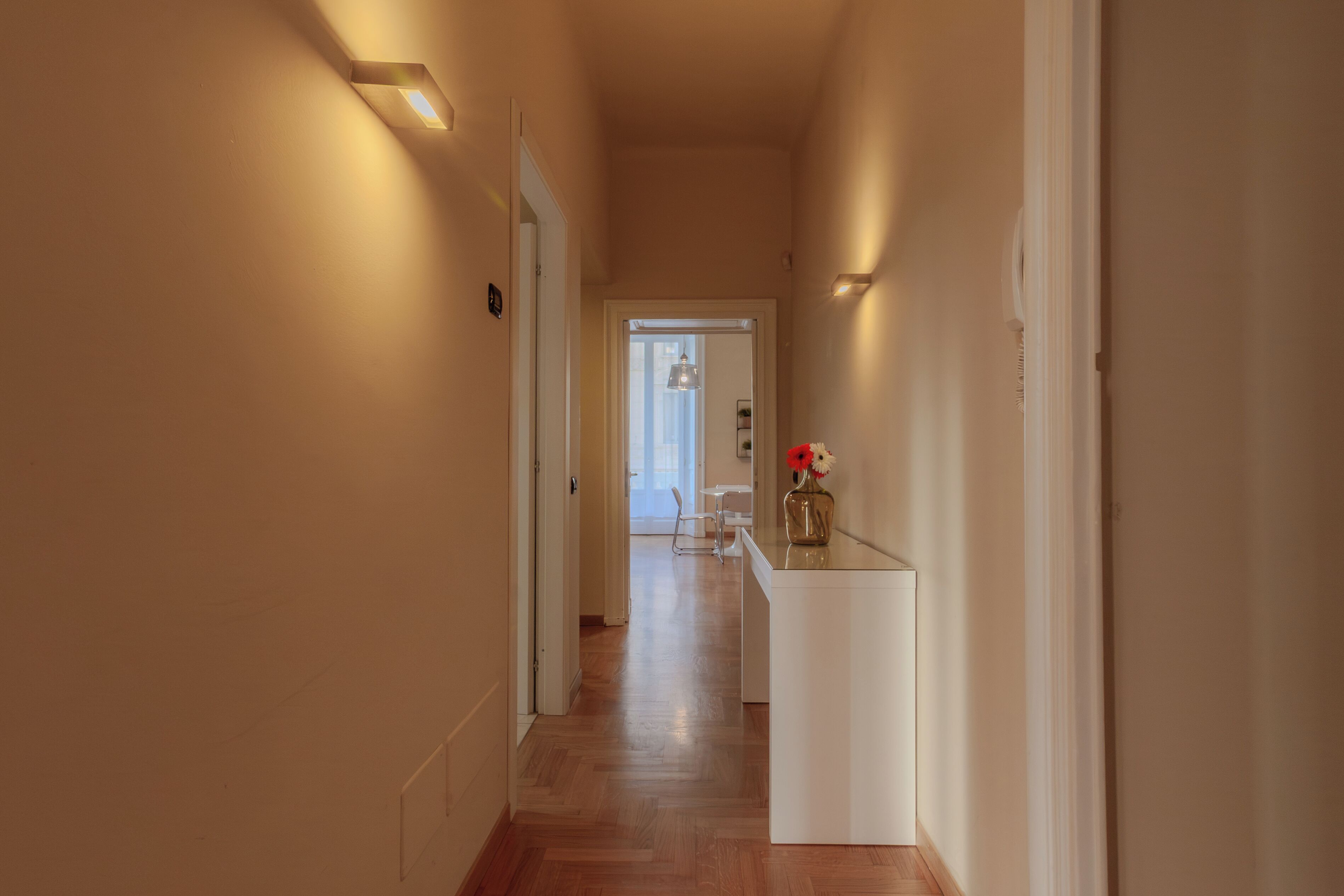 Un couloir doté d’un plancher en bois, de murs blancs et d’une petite table sur laquelle est posé un vase de fleurs.