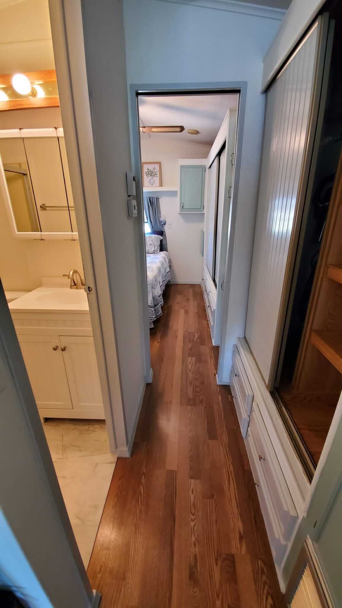A narrow hallway with wooden flooring, a bed with a patterned bedspread, and a window with blinds.