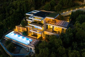 Ett modernt hus med flera våningar, ett poolområde på taket, solpaneler och en välskött trädgård.