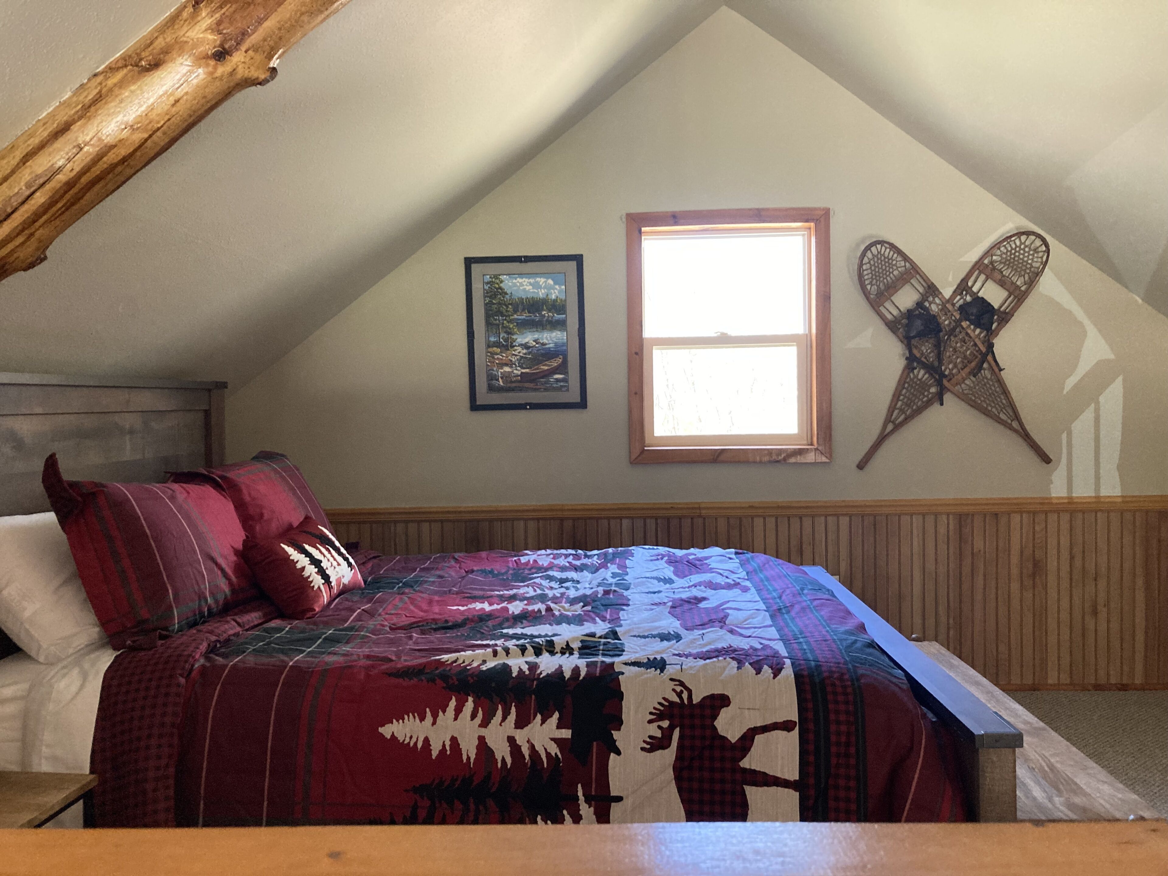 A bedroom with a sloped ceiling, a bed with a red and black patterned bedspread, a framed picture, a window, and a woven wall hanging.