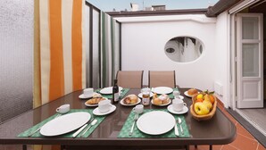 A dining table set with plates, cups, and a fruit bowl, surrounded by chairs in an outdoor patio area with a view of the sky and a building in the background.