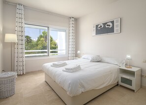 Un dormitorio con una cama grande, una mesita de noche con lámpara, una obra de arte en la pared y vistas al verdor exterior.