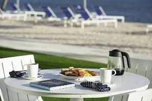 A table with breakfast items, a book, and a coffee maker on a white table outdoors with beach chairs in the background. - Club Suites at Rum Point (North Side)