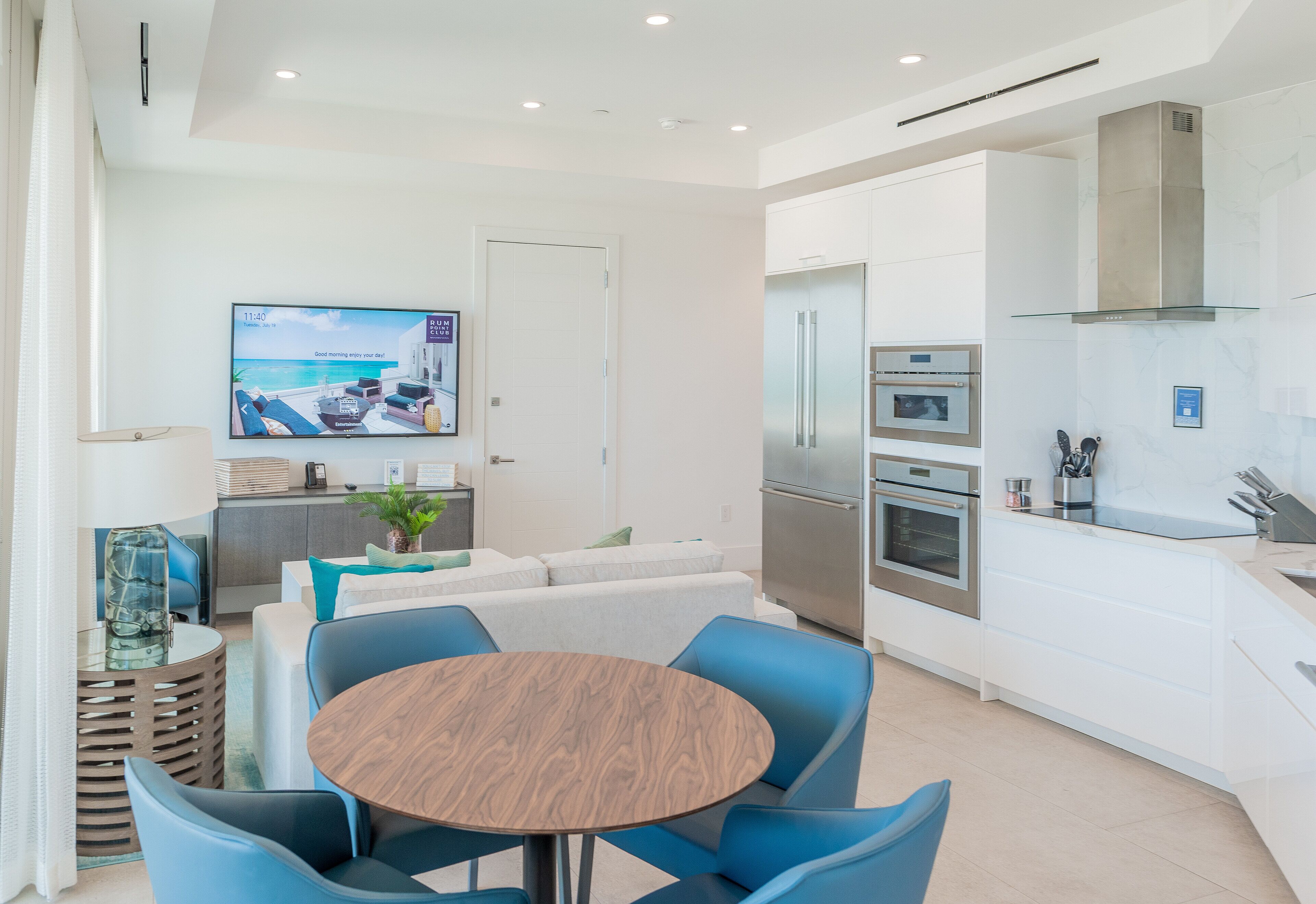 A modern kitchen with a built-in oven, a refrigerator, and a glass-top dining table with blue chairs.