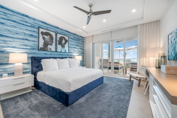 A modern bedroom with a large bed, a ceiling fan, and a view of the outdoors through a sliding glass door. - Club Suites at Rum Point (North Side)