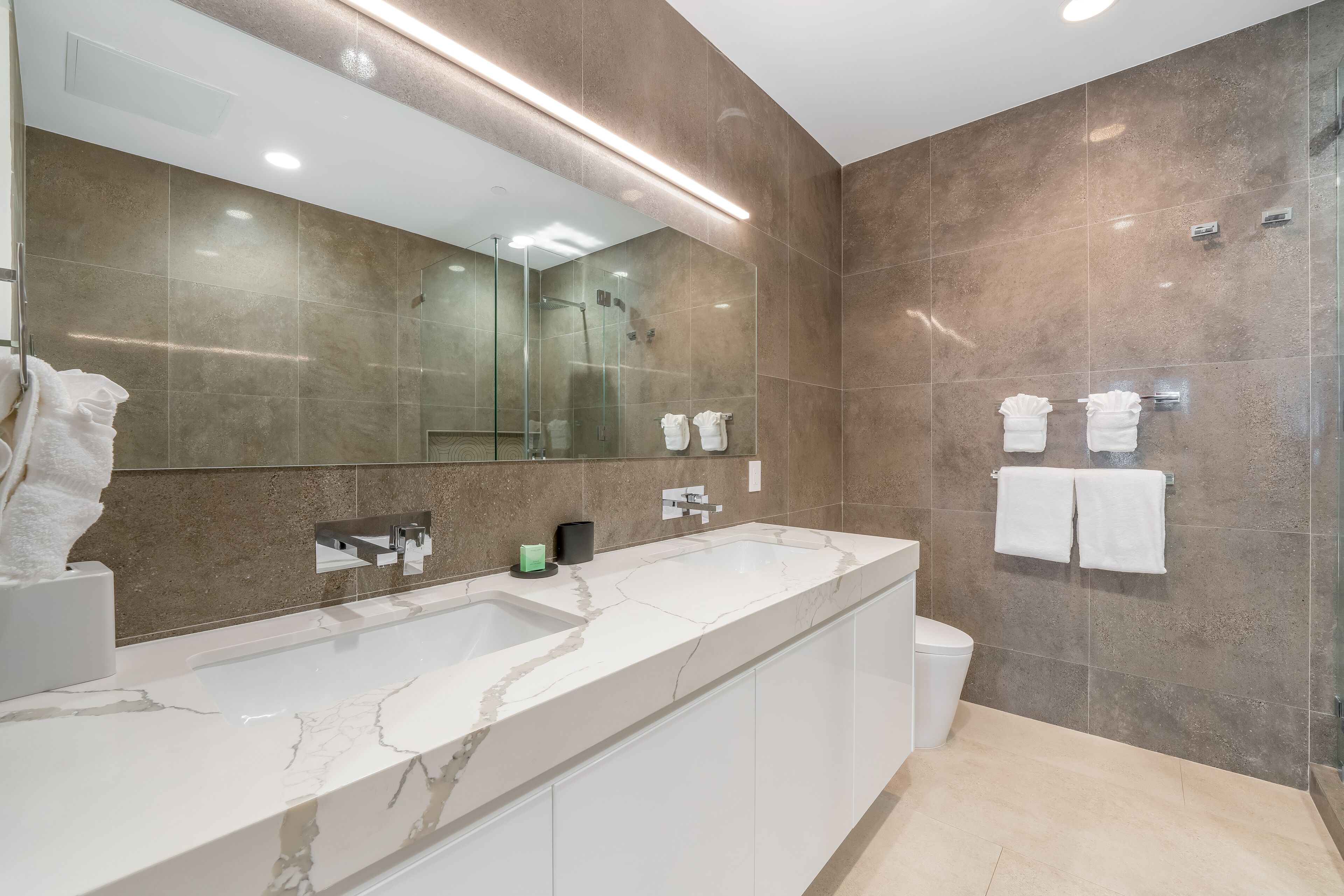 Un baño moderno con un espejo grande, ducha con mampara de vidrio y lavamanos de mármol.
