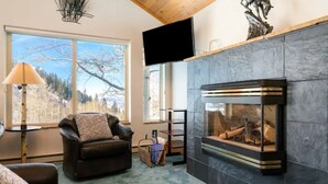 A living room with a fireplace, a leather sofa, a television mounted on the wall, and a large window with a view of trees. - Close to town cabin w/Hot Tub in Steamboat Springs (Steamboat Springs)