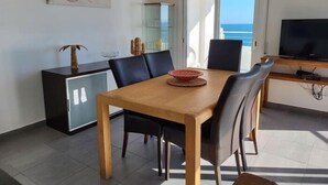 Ein Essbereich mit Holztisch und -stühlen, einer Glastruhe, einem Fernseher auf einem Holzschrank und Blick auf das Meer durch ein großes Fenster.