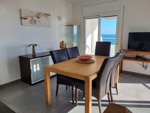 A dining area with a wooden table and chairs, a glass cabinet, a TV on a wooden stand, and a view of the sea through a large window.