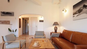 Een woonkamer met een bruine leren bank, twee grijze fauteuils, een houten bijzettafeltje met een hoed, bril en kom, en een lamp.