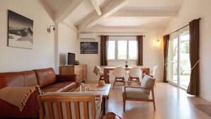Een woonkamer met een bruine leren bank, houten stoelen, een houten salontafel en een televisie op een houten dressoir.