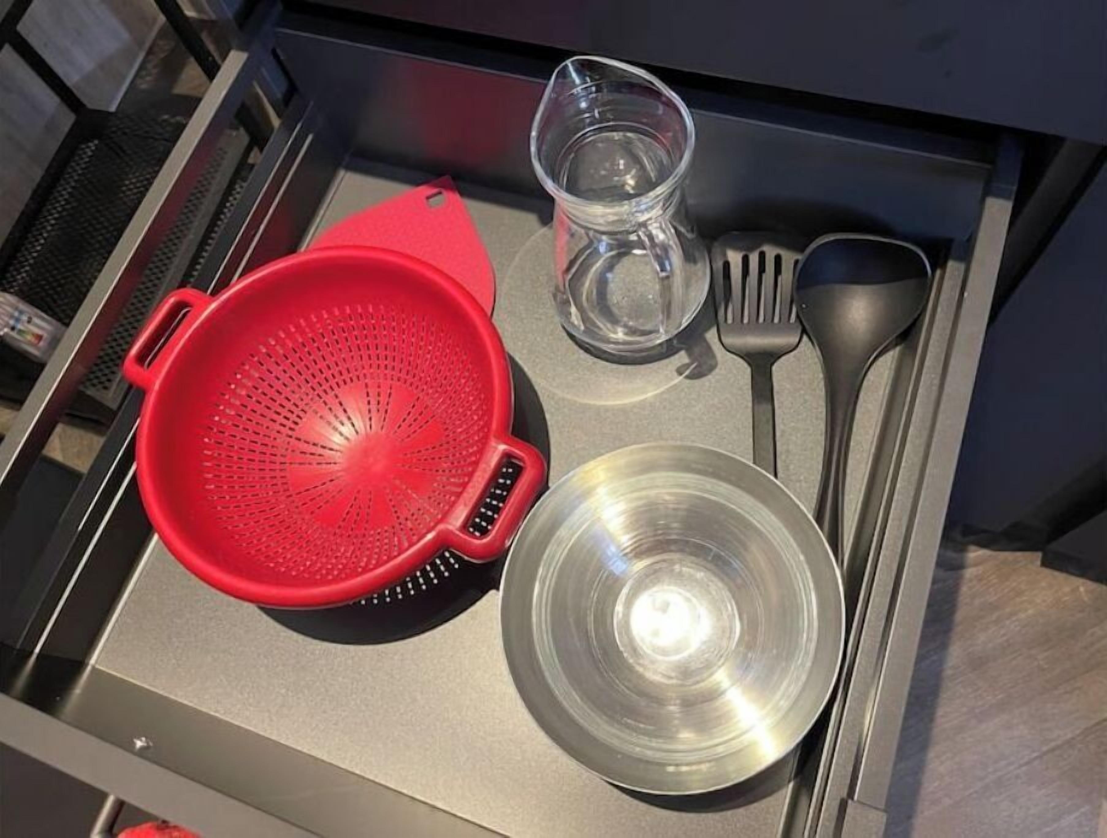 A kitchen drawer with a red colander, a clear glass pitcher, a metal spatula, and a metal bowl.