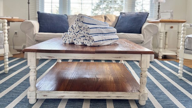 Une table basse rustique en bois, agrémentée de serviettes roulées, se trouve dans une pièce lumineuse et aérée, avec un tapis à rayures, des meubles blancs et une vue dégagée à travers de grandes fenêtres.