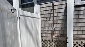 Une terrasse en bois peinte en blanc mène à un espace de douche extérieur abrité, équipé d’un tuyau et d’un pommeau de douche en métal.