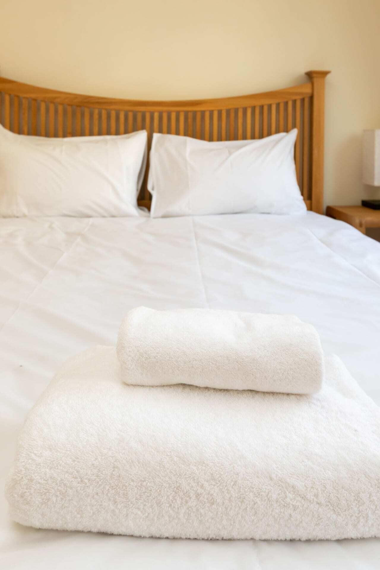 A neatly made bed with white linens and pillows, a wooden headboard, and a folded white towel on the bed.