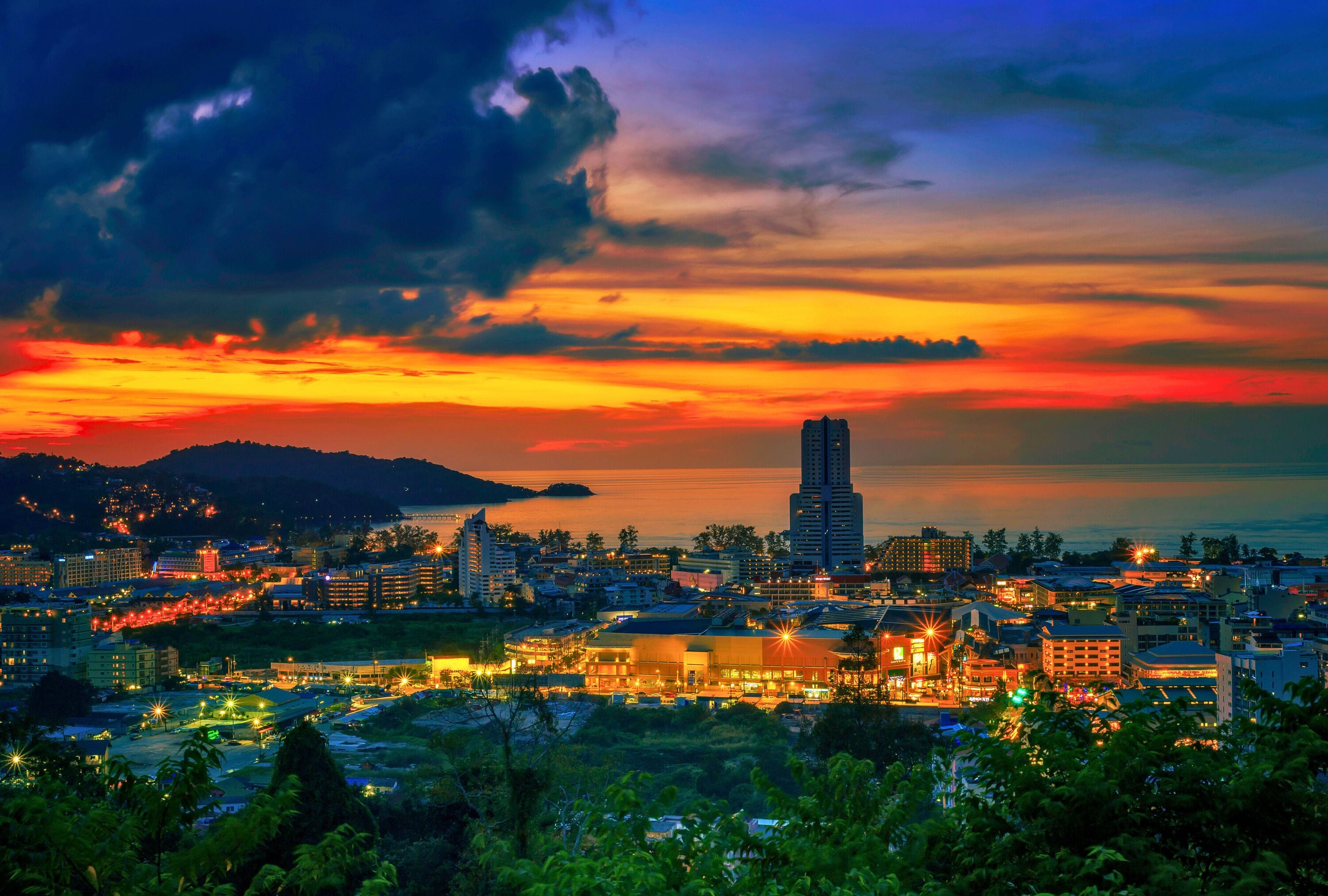A cityscape at sunset with a prominent skyscraper, various buildings, and a body of water in the distance.