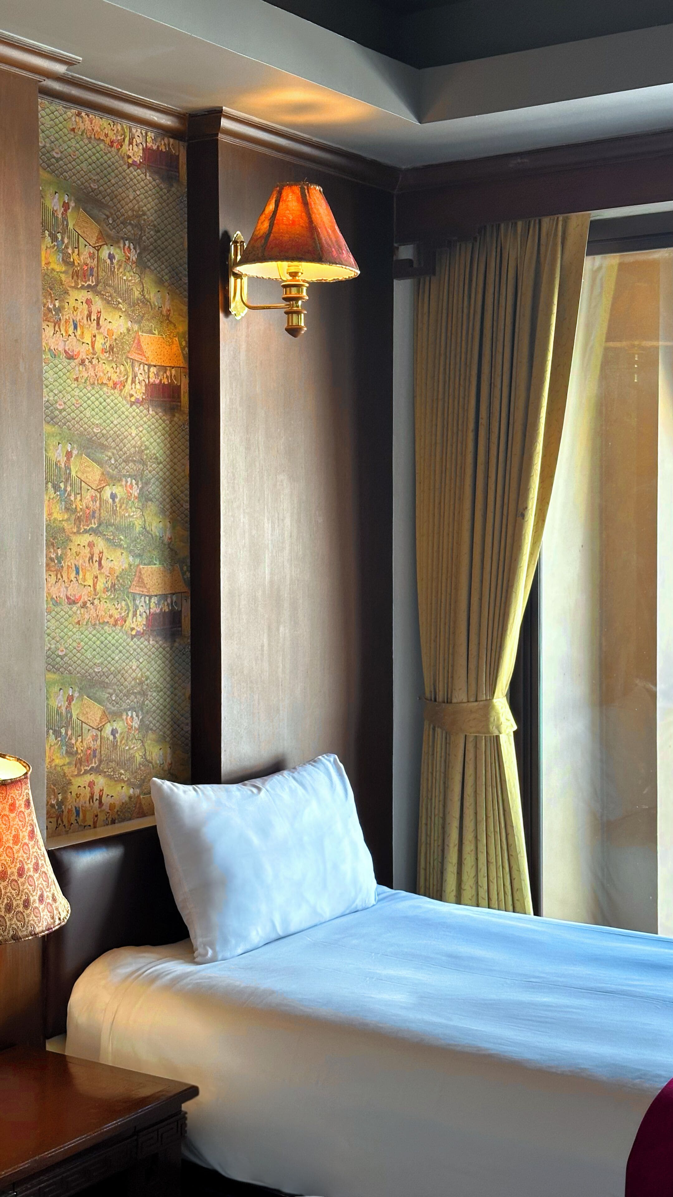 A hotel room with a bed, a wall-mounted lamp, a large window with curtains, and a decorative wall hanging.