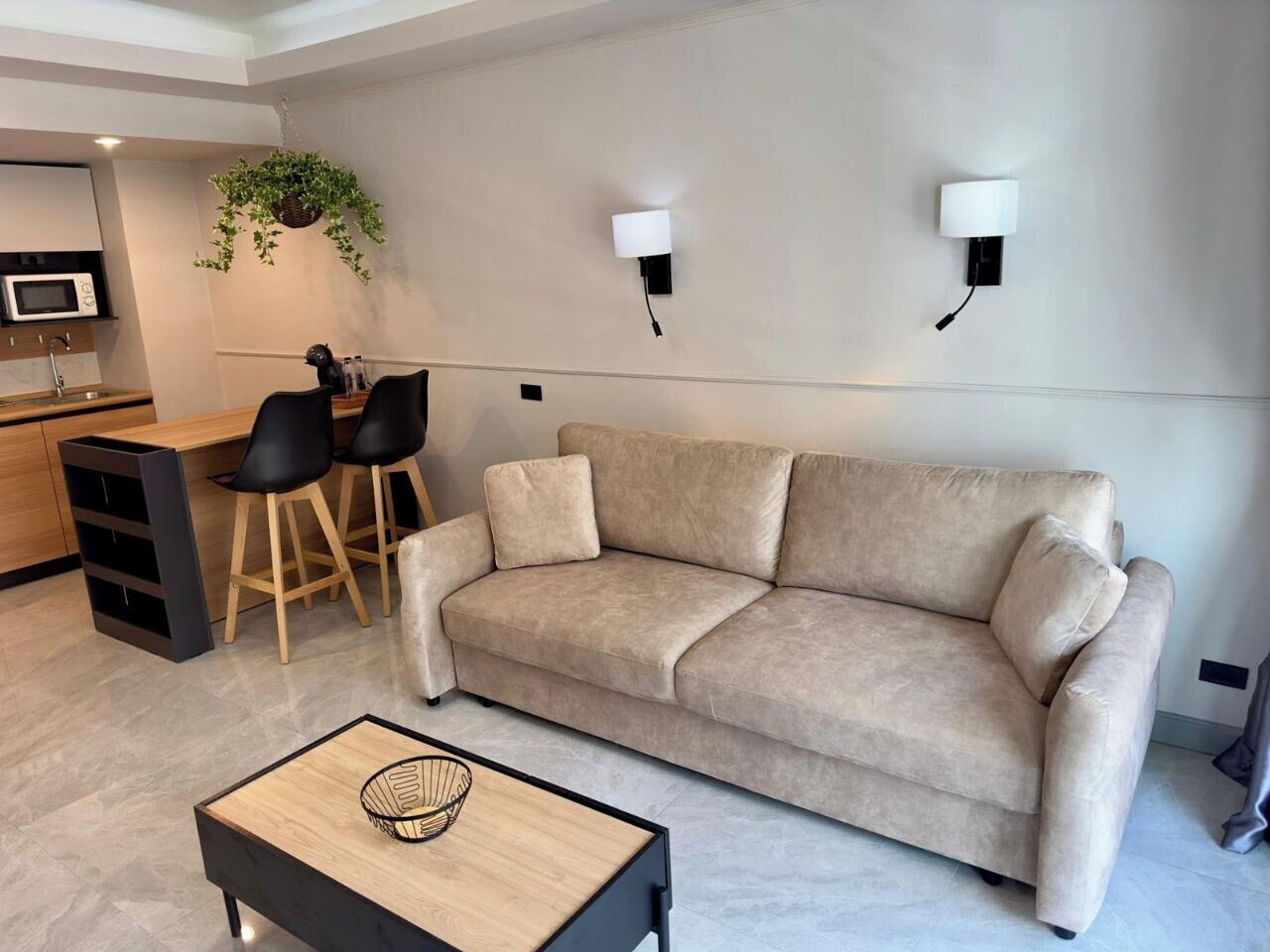 A modern living room with a beige sofa, two black chairs, a wooden coffee table, and a hanging plant.