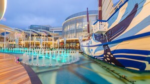 Cubierta de crucero con una piscina grande, un edificio moderno con ventanales y un barco con franjas azules y blancas.