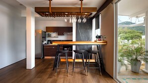 Una cocina moderna con encimera de madera, tres taburetes negros y un amplio ventanal con vistas a la vegetación.