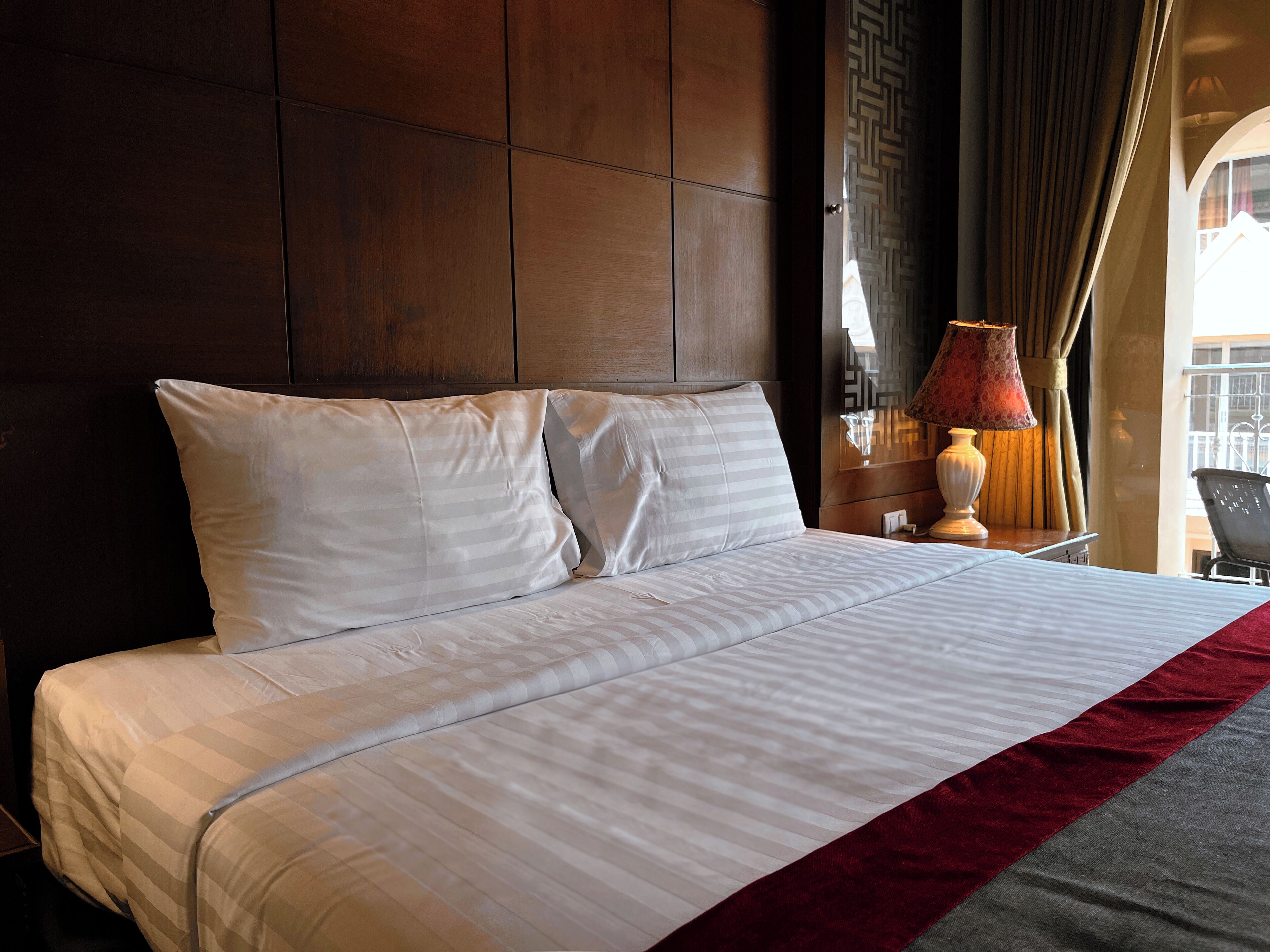 Quarto de hotel com uma cama grande, cabeceira de madeira, mesa de cabeceira com abajur e vista para o exterior pela janela.