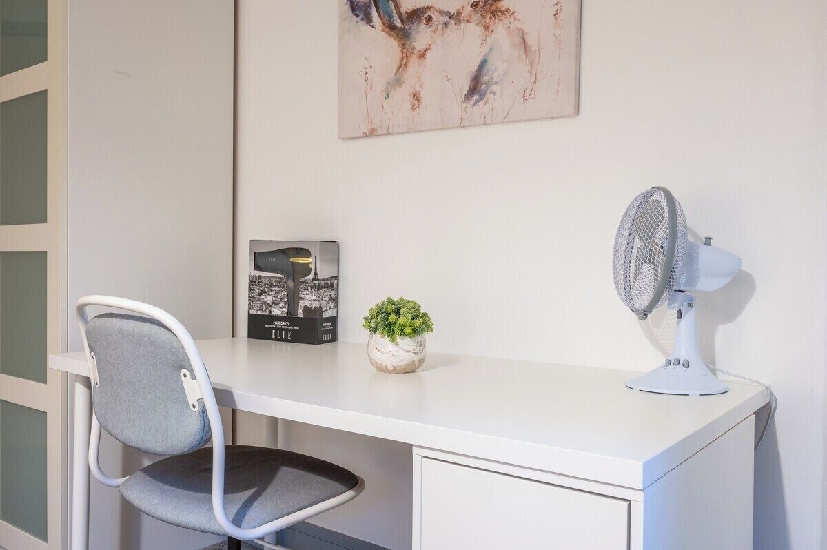 A white desk with a chair, a small potted plant, a framed picture, and a white standing fan.