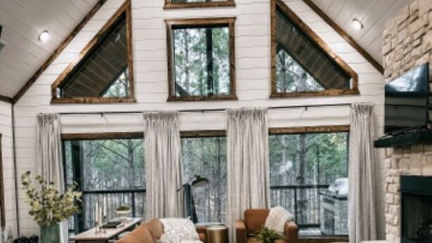 A spacious loft-style living area with a high ceiling, large windows, and a stone fireplace. - Dreamcatcher Cottage- A Luxurious Romantic Escape in Hochatown (Broken Bow)