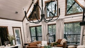 A spacious living room with a large wooden dining table, white upholstered chairs, and a cozy seating area with a brown sofa and a white armchair. - Dreamcatcher Cottage- A Luxurious Romantic Escape in Hochatown (Broken Bow)