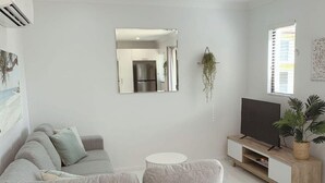 Un salon moderne avec un canapé gris, une petite table blanche, un téléviseur posé sur un meuble en bois et un miroir accroché au mur.