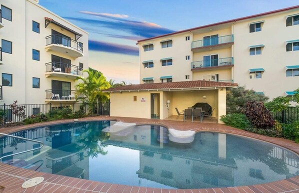 Un espace piscine avec un ciel dégagé, un petit bâtiment doté d’une terrasse, et un immeuble à plusieurs étages avec des balcons.