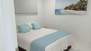 Une chambre à coucher avec un lit, deux oreillers, une couverture bleue, une fenêtre avec des stores et un tableau représentant une scène de plage accroché au mur.