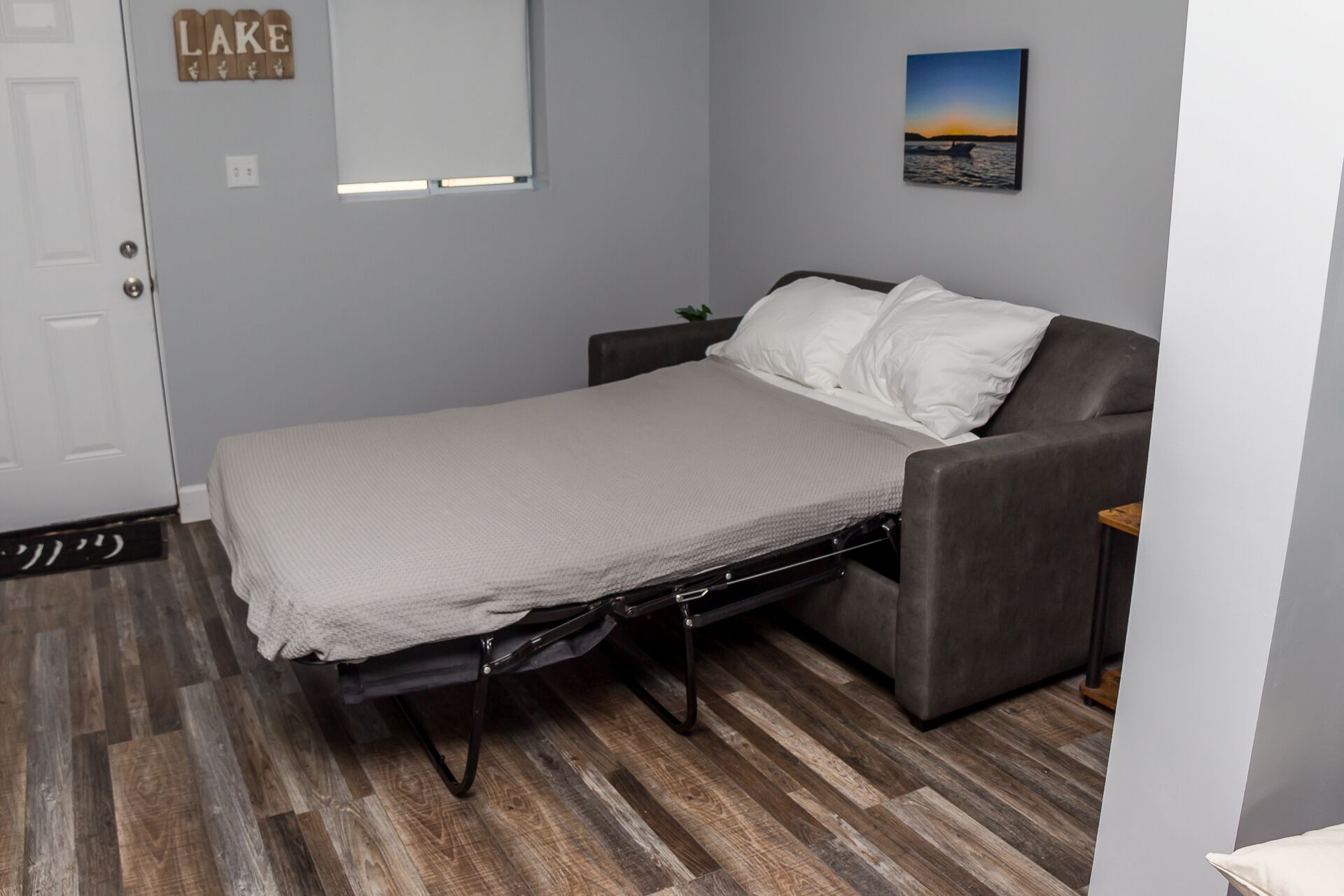 A compact bedroom with a bed, a grey sofa, a small table, and a wall-mounted picture.