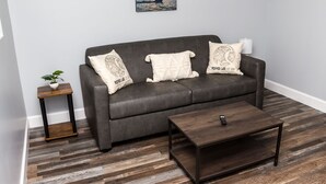 A grey sofa with three decorative pillows, a small wooden side table with a plant, and a dark wooden coffee table in a living room with wooden flooring.