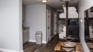 A compact hotel room with bunk beds, a small kitchenette, a trash bin, and a dining area with a table and chairs.